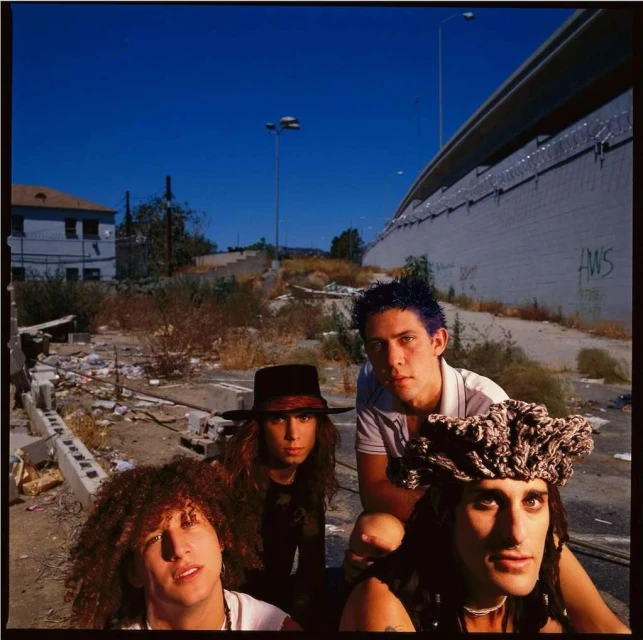 Four band members with curly hair and hats pose outdoors in a gritty urban setting with street art, rubble, and an overpass above them.