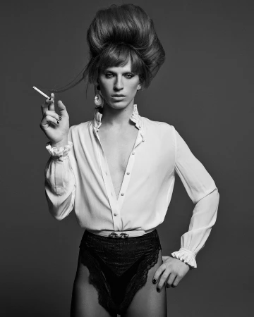 Portrait of a person with large teased beehive hair wearing a ruffled white shirt and holding a cigarette, shot by Mark Seliger.