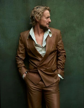 Owen Wilson wearing a brown leather suit and striped shirt against a dark green textured background in a portrait by Mark Seliger