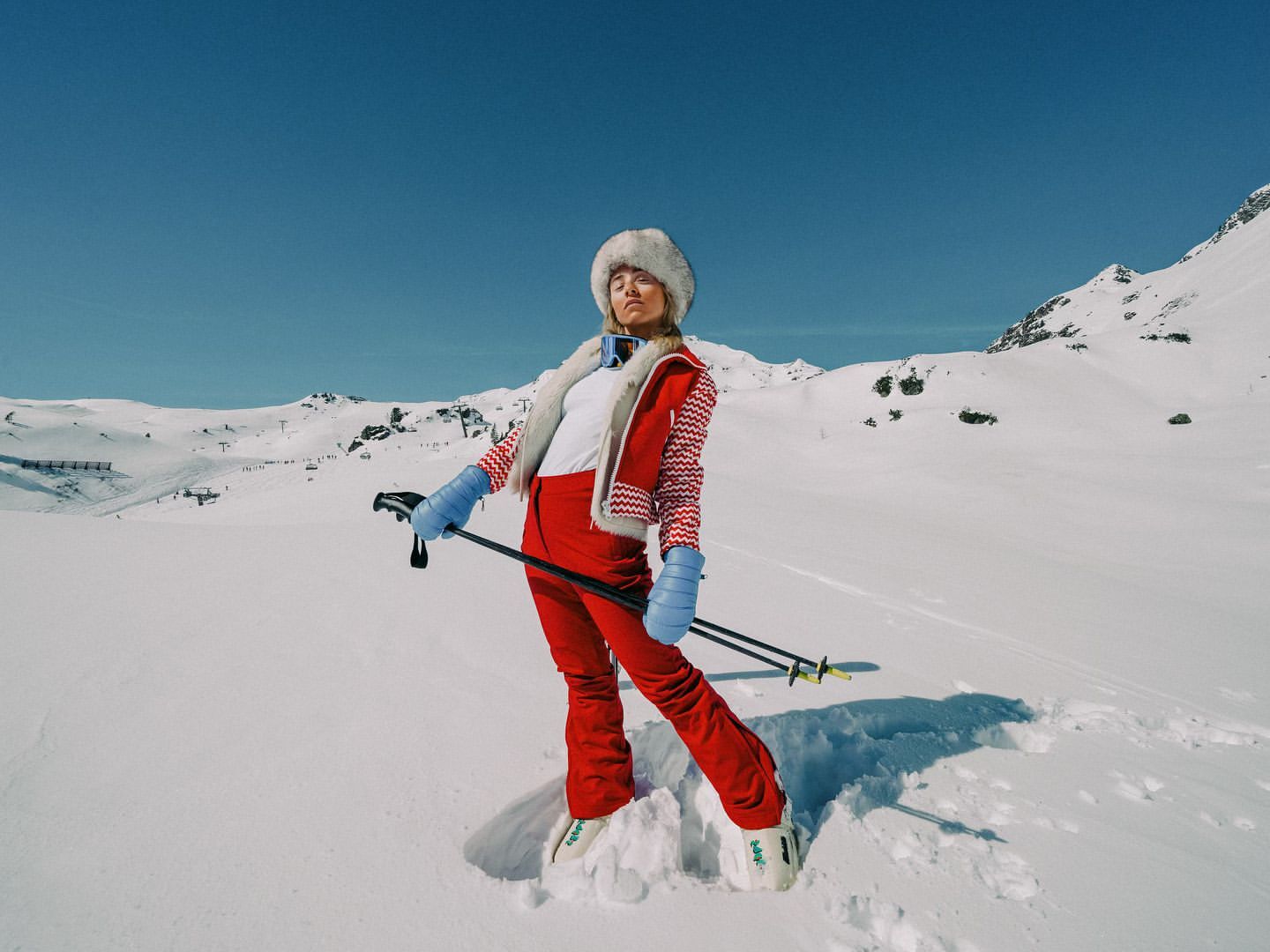 A woman in red ski suit and fur hat poses in the snow with ski poles under a clear blue sky, surrounded by a mountain range.