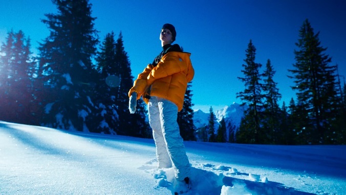 Adult standing in a snowy winter landscape featuring mountain peaks and evergreen trees under a clear blue sky wearing outdoor gear.