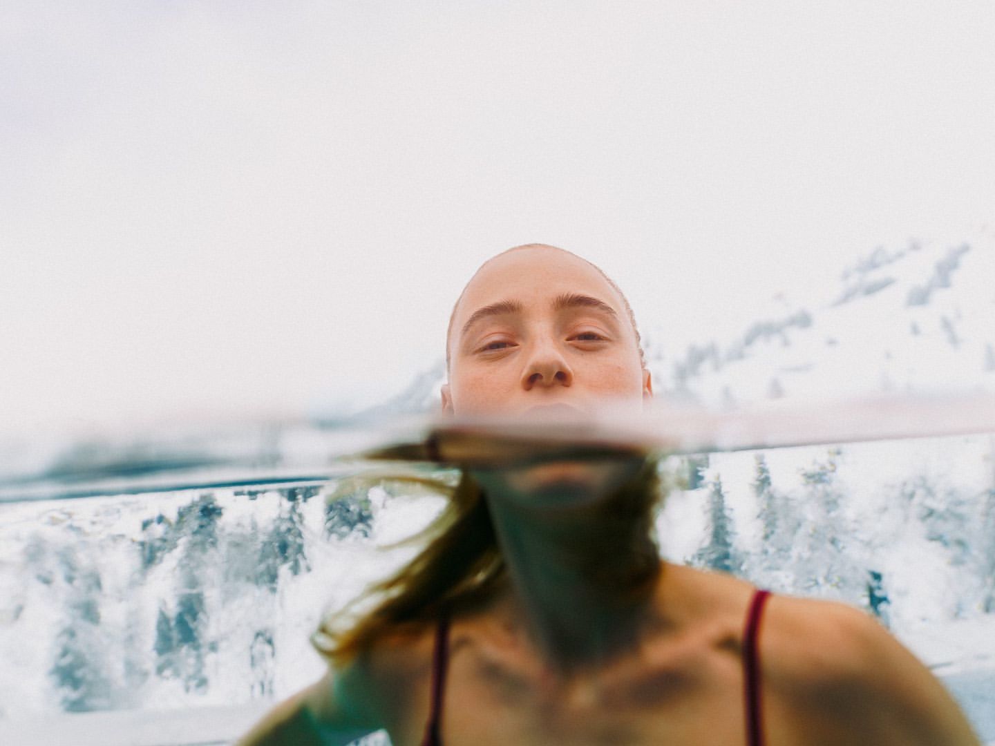 Woman in swimsuit underwater, viewed from chest-up, with snowy mountains and overcast sky in the blurred background.