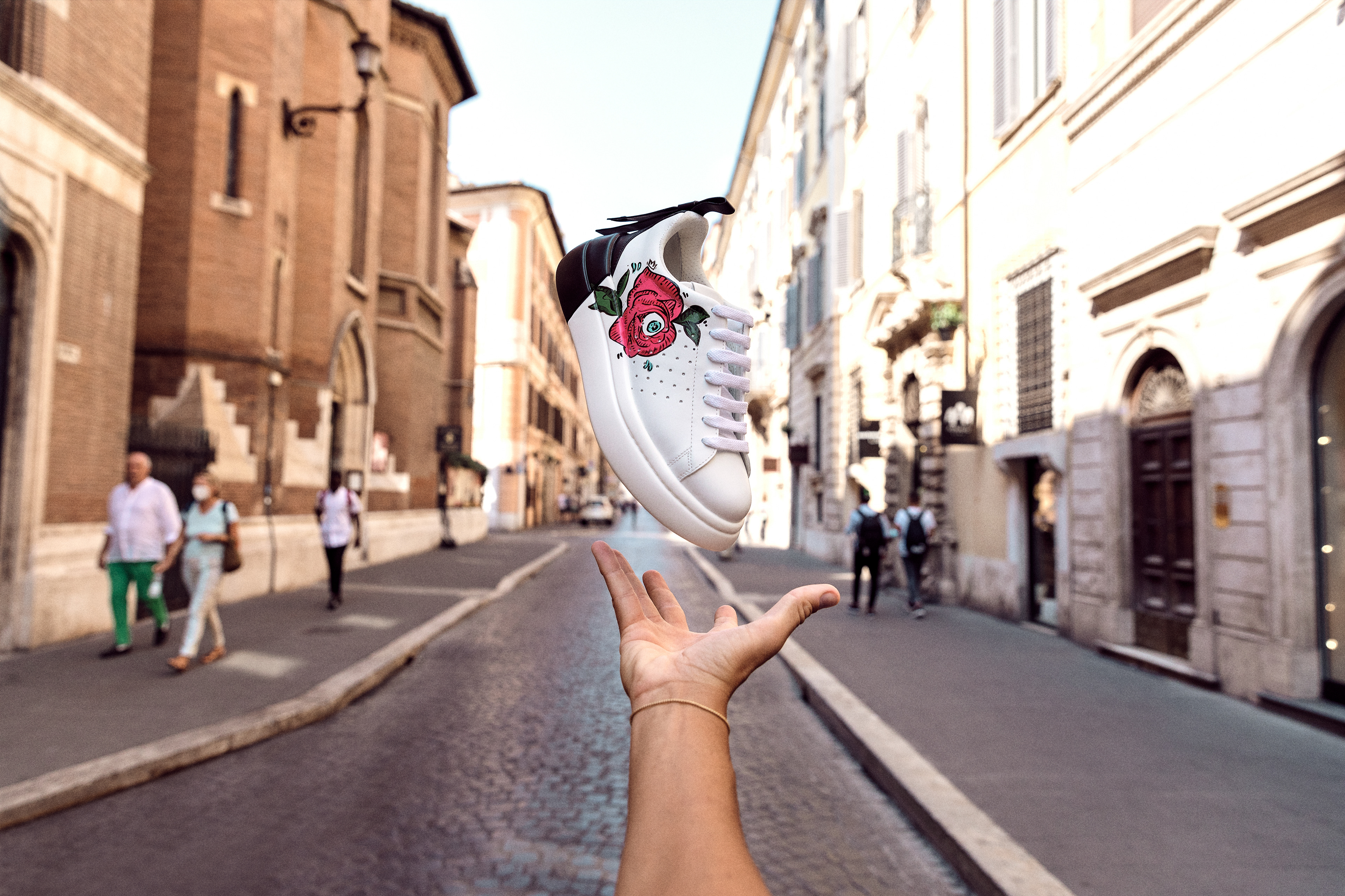 White sneaker with a rose design floating above a hand in an urban environment, featuring blurred pedestrians and buildings.