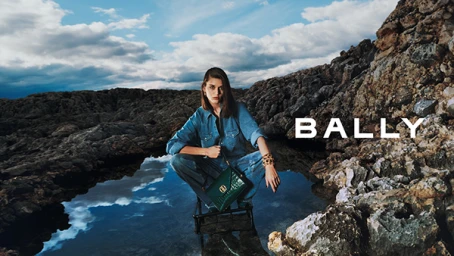 Woman in denim shirt and jeans squatting outdoors near rocky terrain and water holding a green handbag