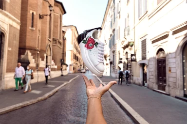 A white sneaker with a red rose graphic floating above an outstretched hand on a historic European street.