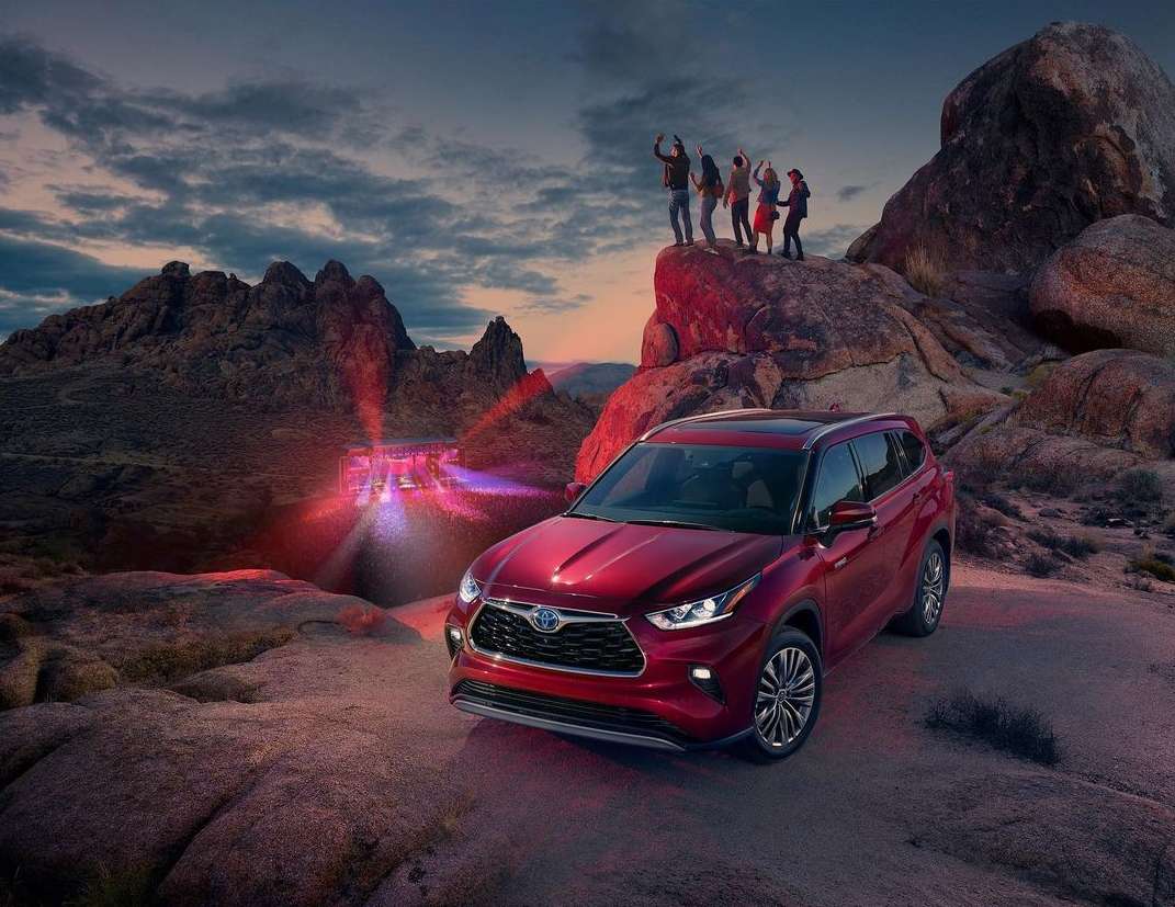 A red Toyota Highlander SUV parked in a desert landscape at dusk, with a group of people on a large rock formation in the background.