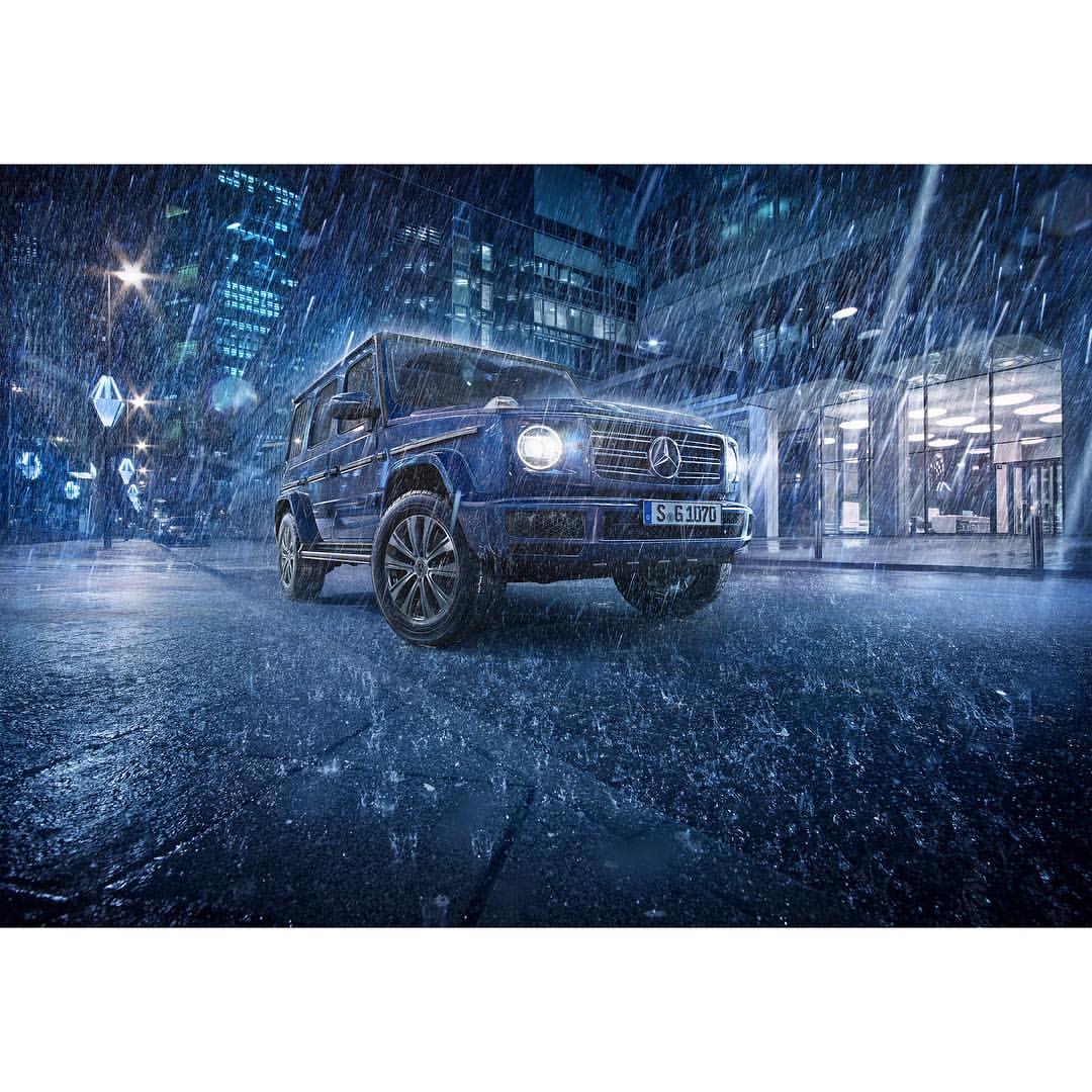 A blue Mercedes-Benz SUV is parked on a wet city street at night in the rain, buildings and streetlights visible in the background.