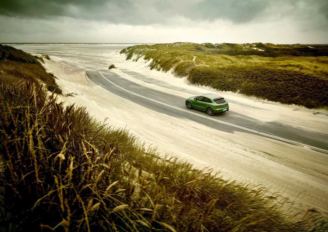 Green SUV driving on asphalt road through a sandy, grassy dune landscape under an overcast sky. Automotive photography.