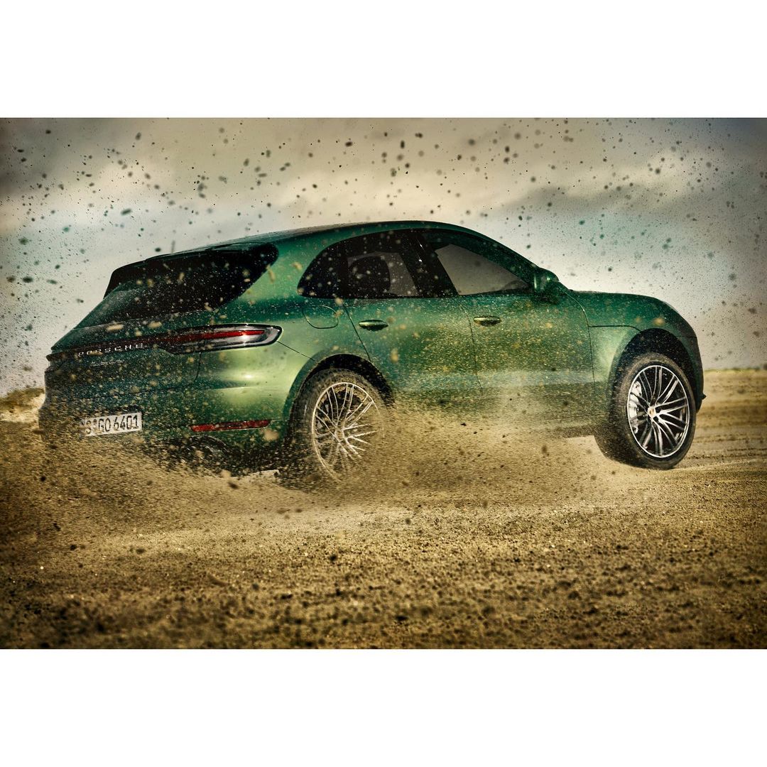 Green Porsche Macan SUV driving on dirt surface with sand splashing from the tires against a cloudy sky, rear view, automotive shot.