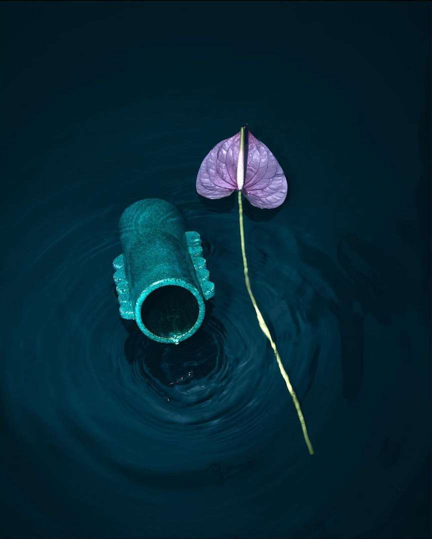 Still life shot of a pink anthurium flower and a green concrete pipe in a dark blue liquid, creating ripples, for surreal, abstract art.