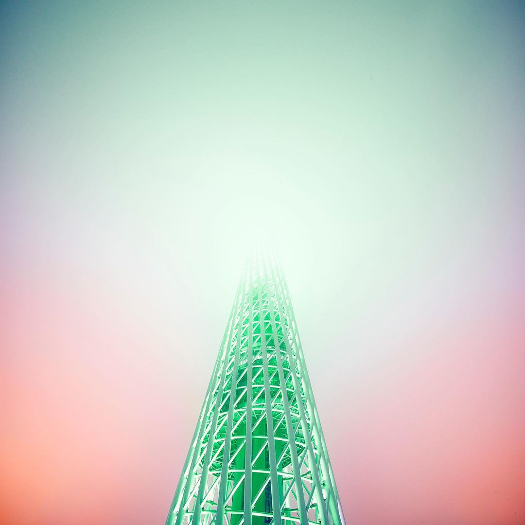 Low angle of a lattice tower disappearing into fog. The structure is green, and the sky is graded from red to green, with fog obscuring the top.