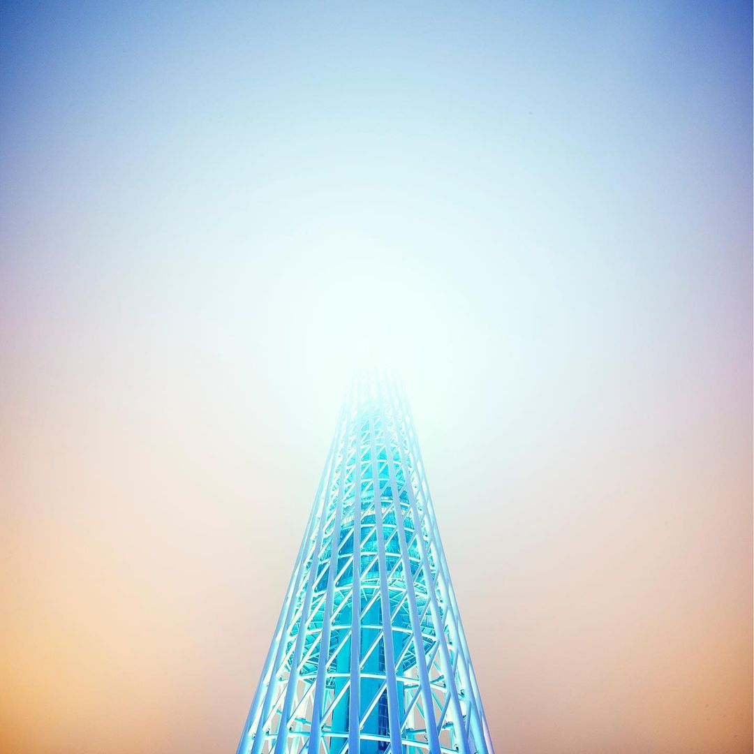 Low-angle shot of a futuristic, blue, steel lattice tower rising through the fog, fading into a bright white sky at daytime.