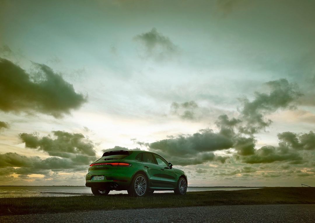 Green Porsche Macan SUV parked on grassy field at sunset with cloudy skies, reflecting luxury and automotive design against a scenic backdrop.