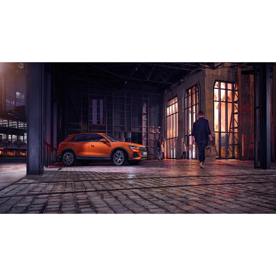 Orange Audi Q2 parked inside a large industrial building, a man walks away from the car, with large windows and industrial structural elements.