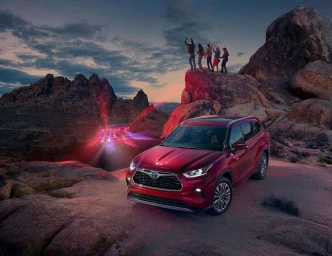 Red SUV parked on rocky terrain at dusk with a group of people standing on a rocky outcrop and distant stage lights.