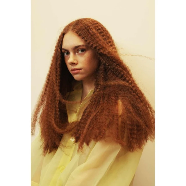 Portrait of a young woman with auburn crimped hair looking at the camera wearing a translucent yellow button-up shirt against a soft, muted yellow background.