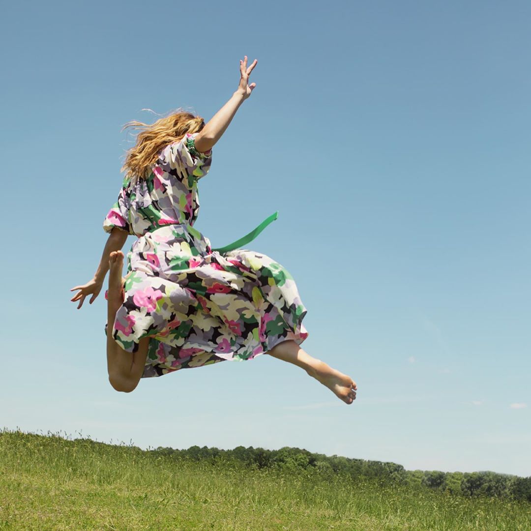 Adult woman barefoot jumping in a floral dress in a grassy field under a clear blue sky, with a green belt, showcasing dynamic movement.