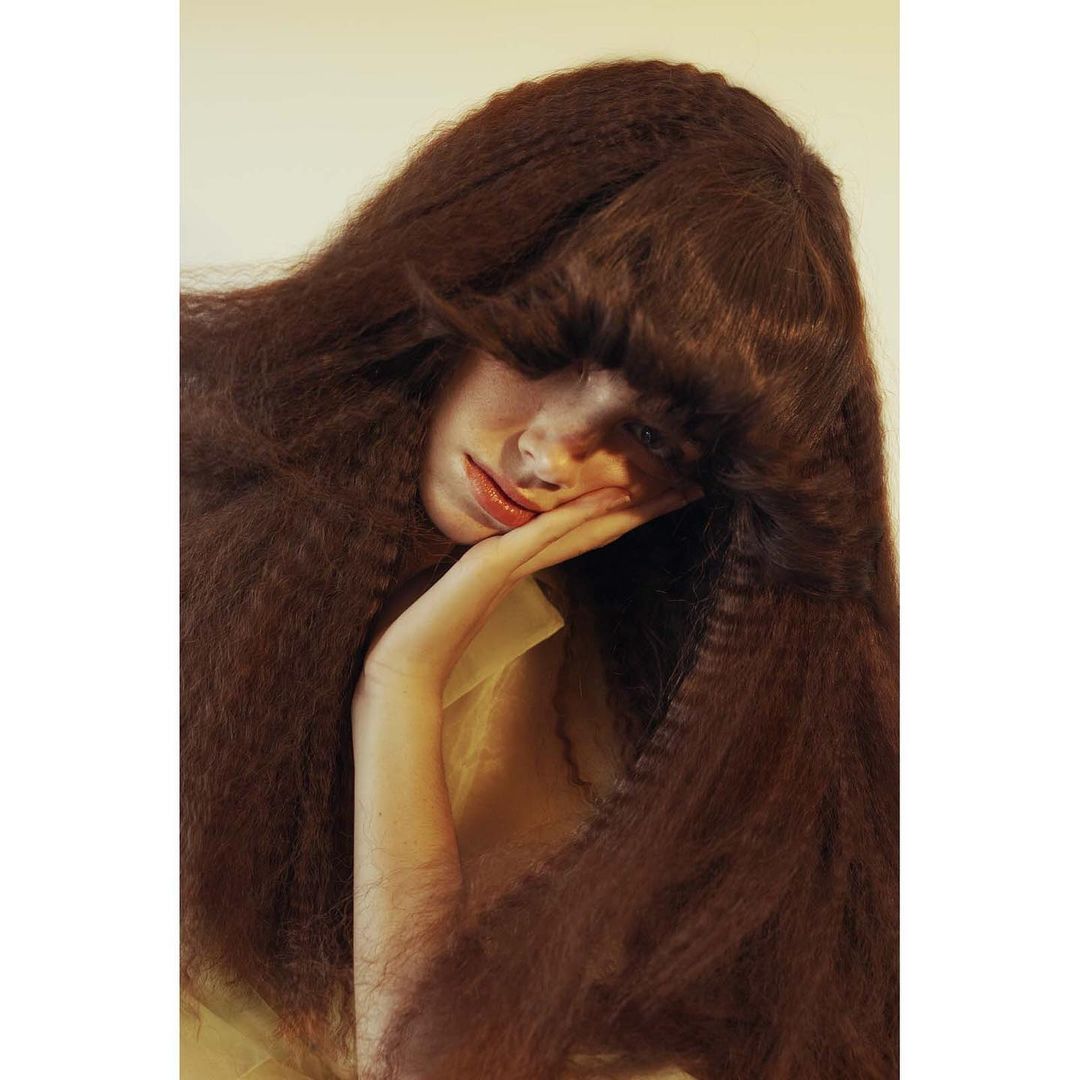 Portrait of a young woman with long crimped auburn hair resting her face on her hand in a beauty and fashion shot. Indoor lighting.