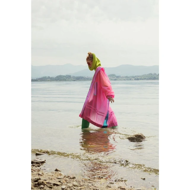 A person in a bright pink transparent coat and green leggings stands in shallow water, near the rocky shoreline of a wide lake.