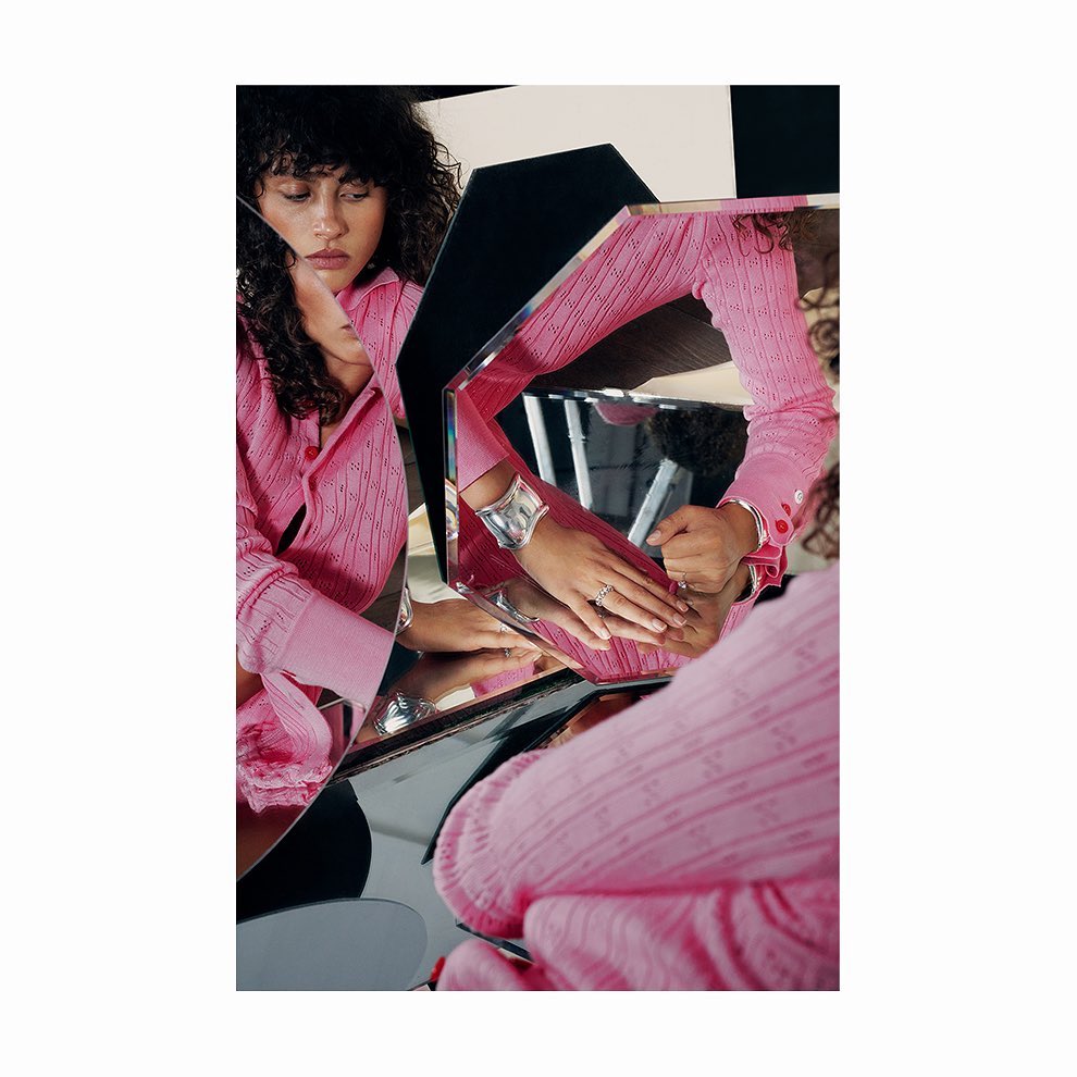 An adult female with curly hair wears a pink button-down shirt and silver bracelet, reflected in geometric mirrors with black & white panels.