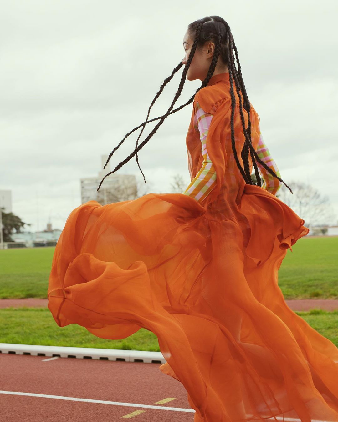 Woman in flowing orange gown poses on running track under overcast sky, fashion editorial with long braids, outdoor setting.