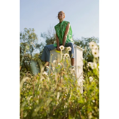 Woman sits on a white box in a field of flowers wearing a green striped blouse and blue pants in a natural outdoor setting.
