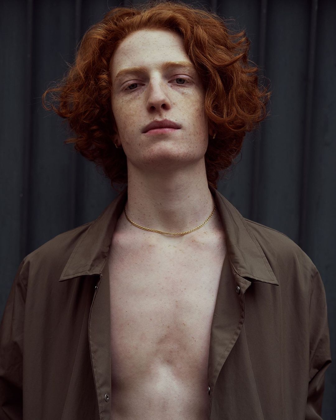 Portrait of a young adult male with red curly hair and freckles wearing an unbuttoned olive green shirt against a dark background.