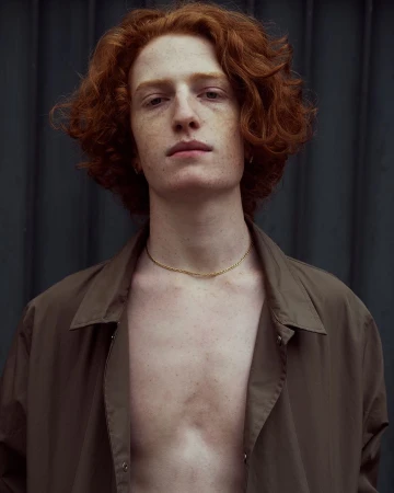 Portrait of a young adult male with red curly hair and freckles wearing an unbuttoned olive green shirt against a dark background.