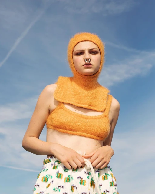 Woman in orange balaclava and crop top poses against blue sky. Summer fashion with floral pants and white rope belt. Outdoor editorial.