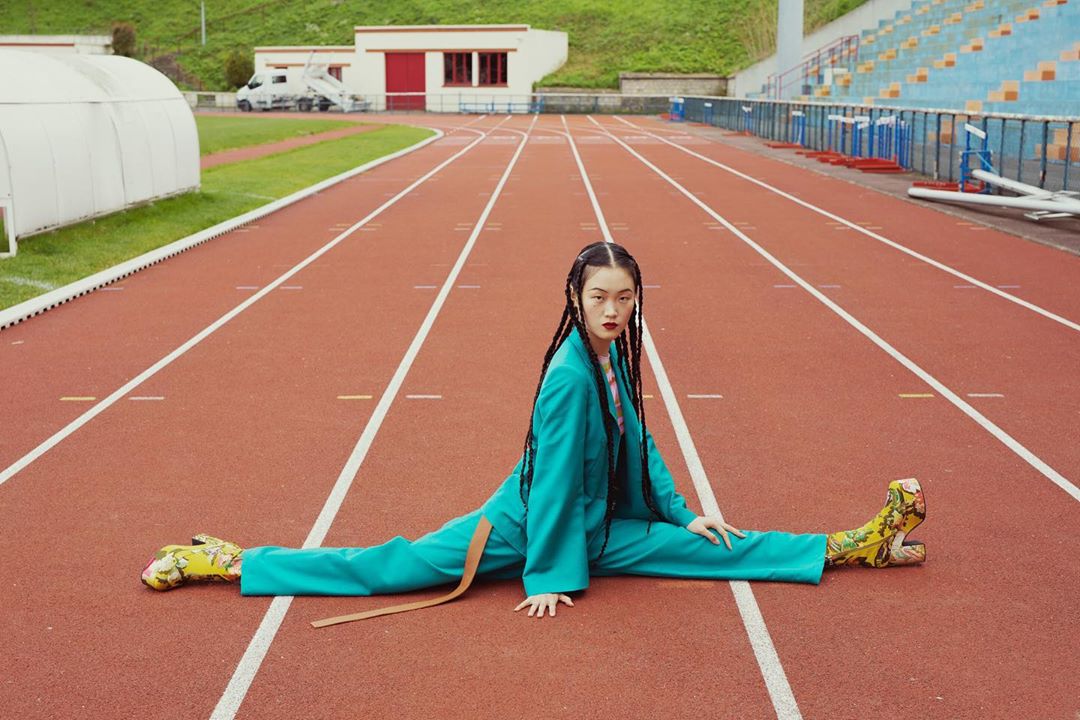 Woman in teal suit and floral boots performs splits on a red running track outside, with a building and trees visible in the background.