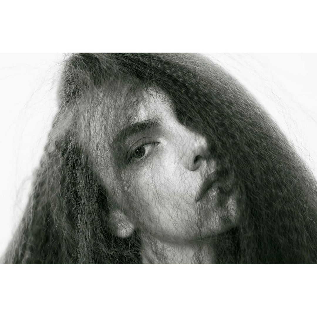 Monochrome portrait of a woman with intricately textured long dark hair framing her face against a stark white backdrop in a beauty shot.