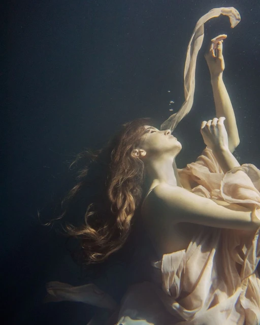 Underwater view of a fair-skinned woman with long, wavy brown hair, wearing a light peach-colored gown, with her arms raised.
