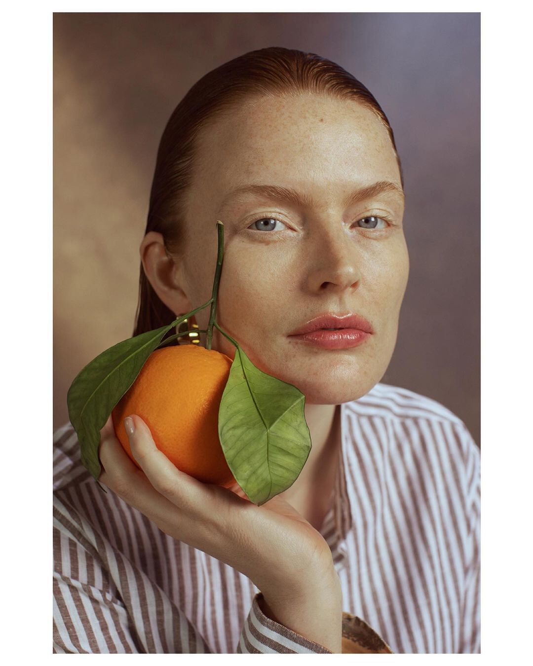 Portrait of a fair-skinned adult woman with wet red hair holding an orange with green leaves. She is wearing a striped shirt and looking at the camera.