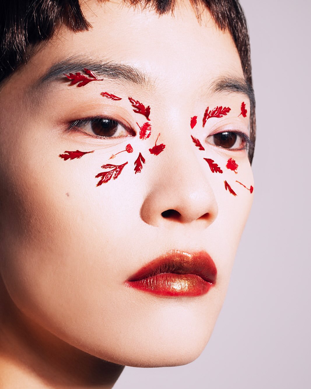 Close-up portrait of an Asian woman with red leaf-shaped face makeup and red lipstick against a neutral grey background.