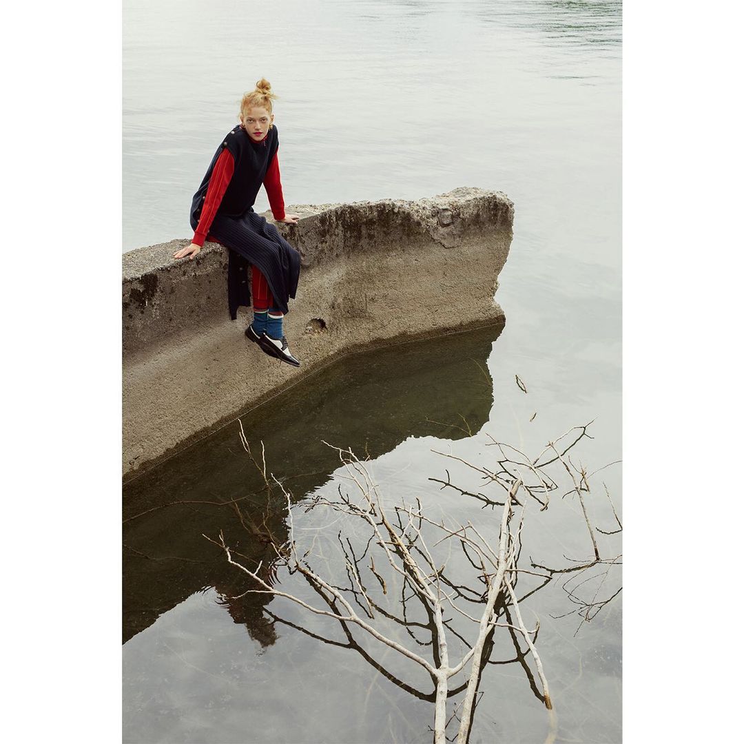 A solo female model with red hair sits on concrete by the water's edge in a long black vest and red top, reflected on the surface.