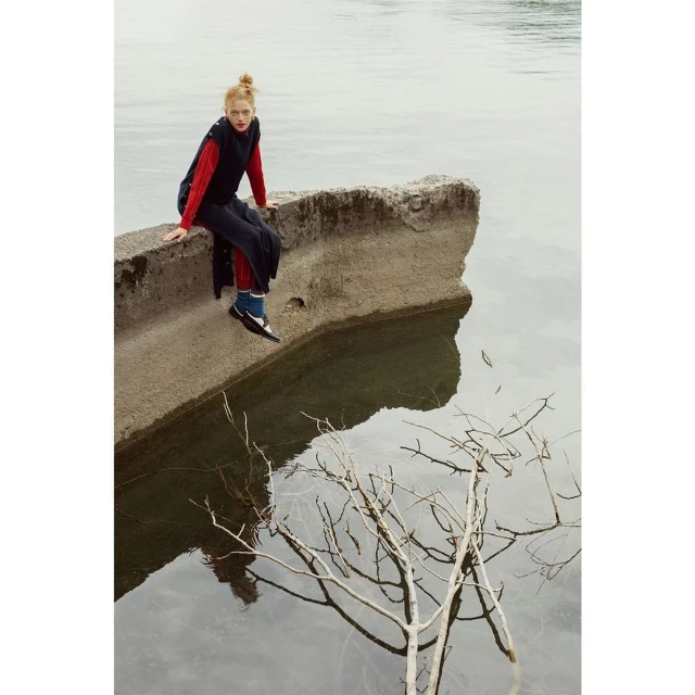 A solo female model with red hair sits on concrete by the water's edge in a long black vest and red top, reflected on the surface.