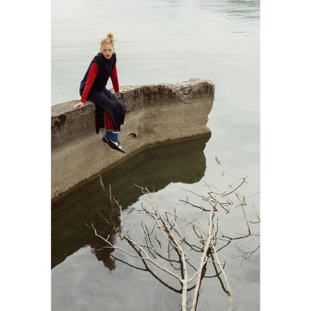 A solo female model with red hair sits on concrete by the water's edge in a long black vest and red top, reflected on the surface.