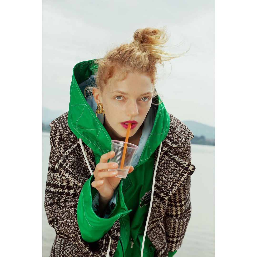 Woman drinks from a clear plastic cup with a straw while wearing a green hooded jacket and patterned coat with an outdoor background.