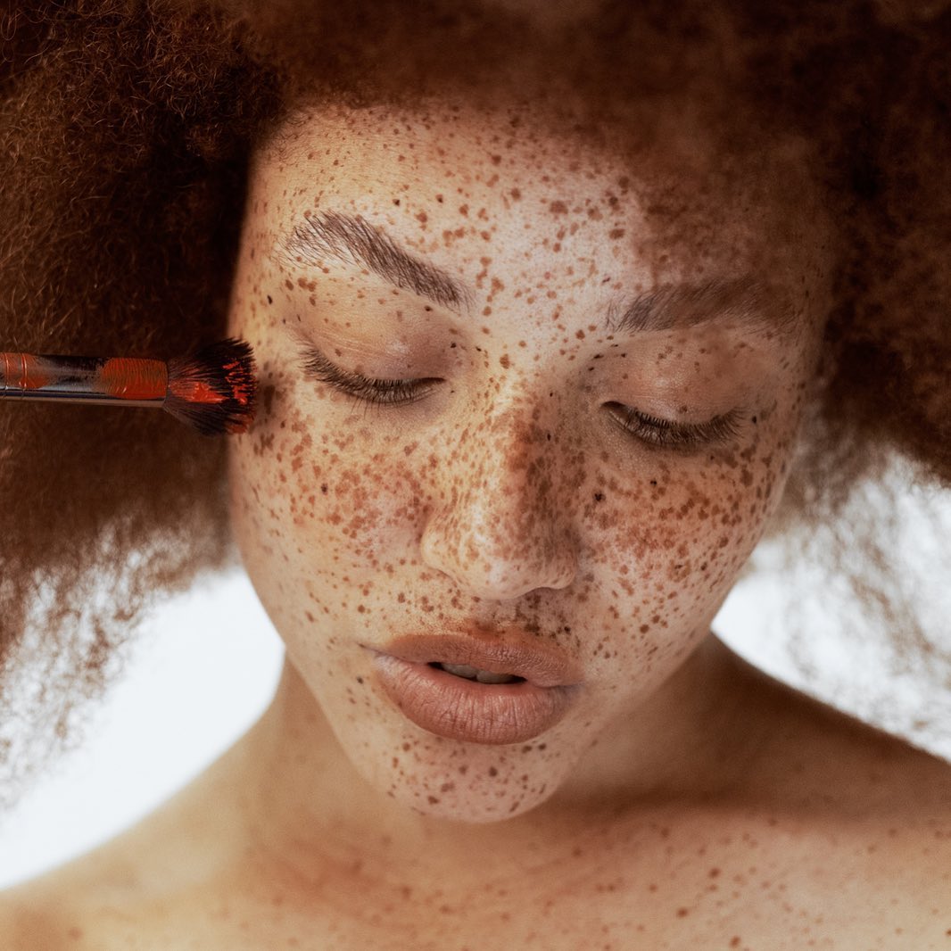 Close-up of a freckled adult woman with closed eyes having makeup applied by a brush on a white background in soft lighting.