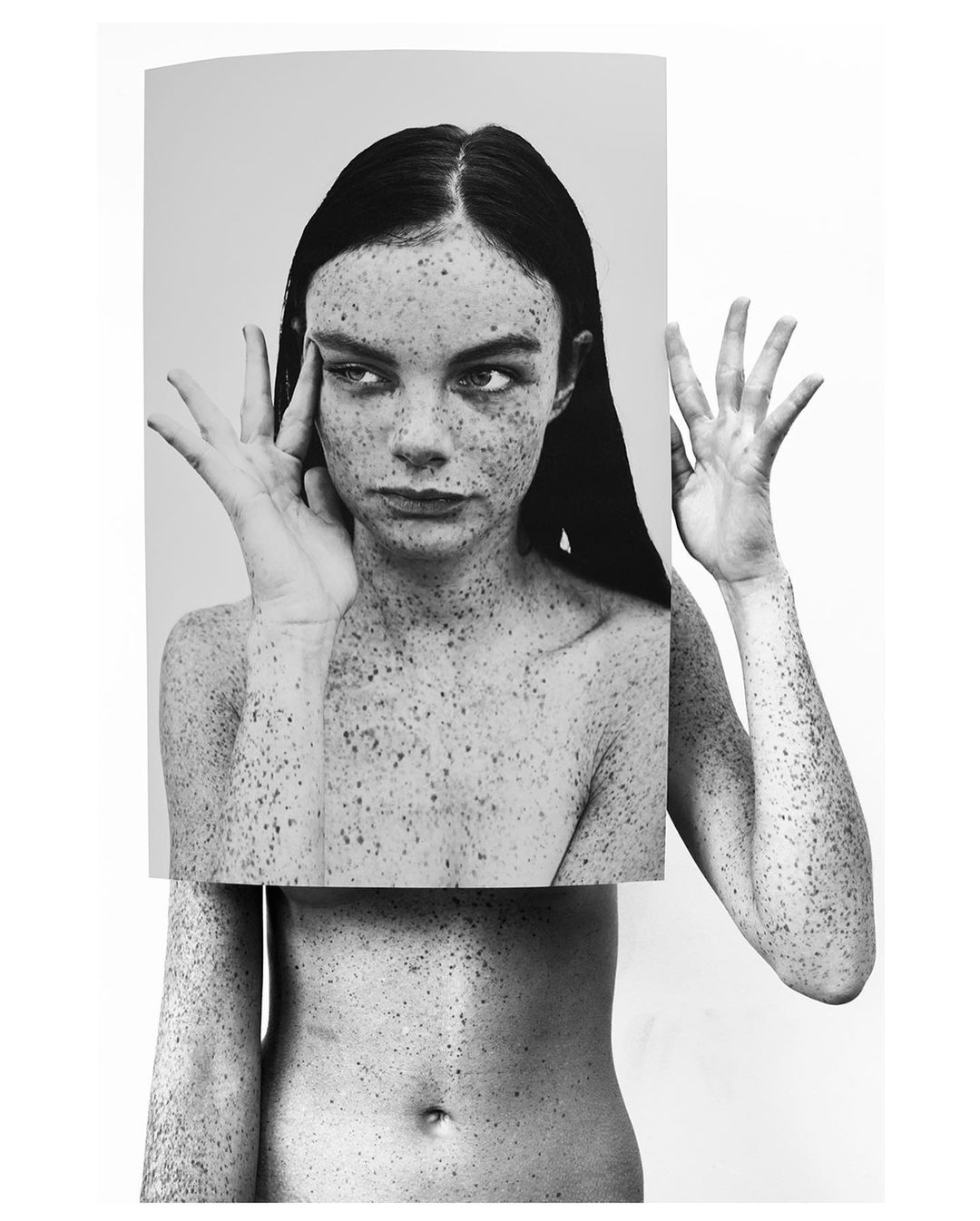 Monochrome portrait of a freckled adult woman holding a print of her face in front of her body, on a white studio backdrop.
