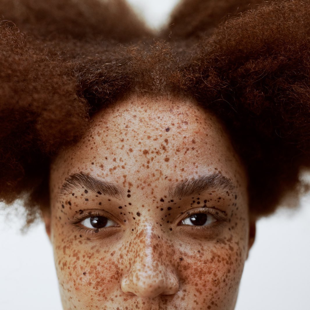 Close-up portrait of an adult female with brown curly hair and freckles on her face, captured in a brightly lit indoor environment.