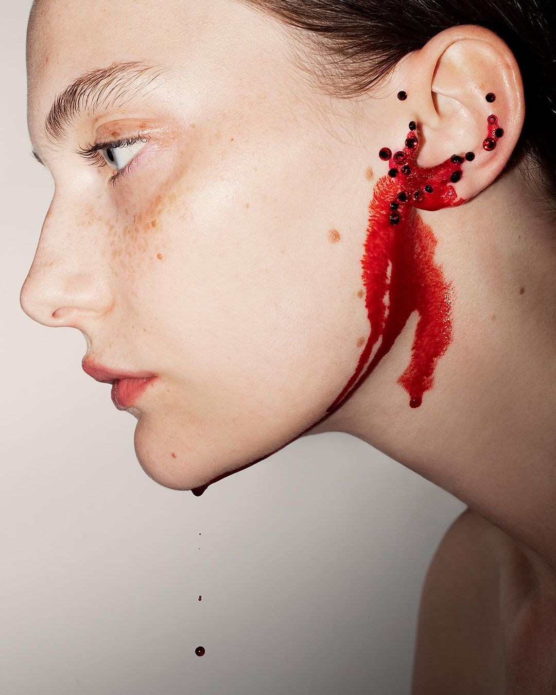 Close-up of a young woman's face in profile with black beads and a red liquid on her ear against a white background.