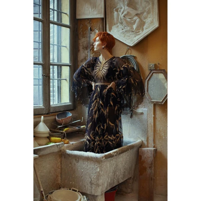 Woman in an ornate dress stands in a vintage tub next to a window, looking out in a well-lit, rustic studio setting with plaster artwork.