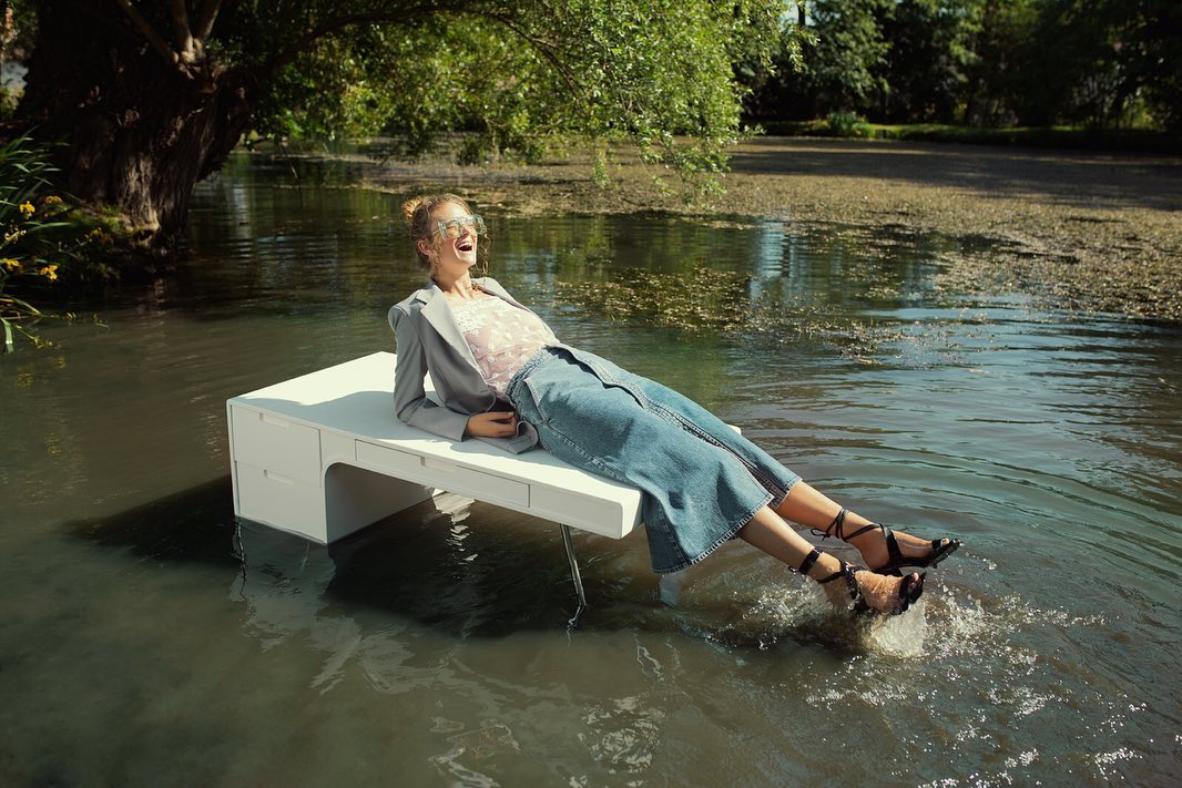 Woman in blazer and skirt lies on white desk floating on lake with trees in background, smiling and splashing feet in the water.