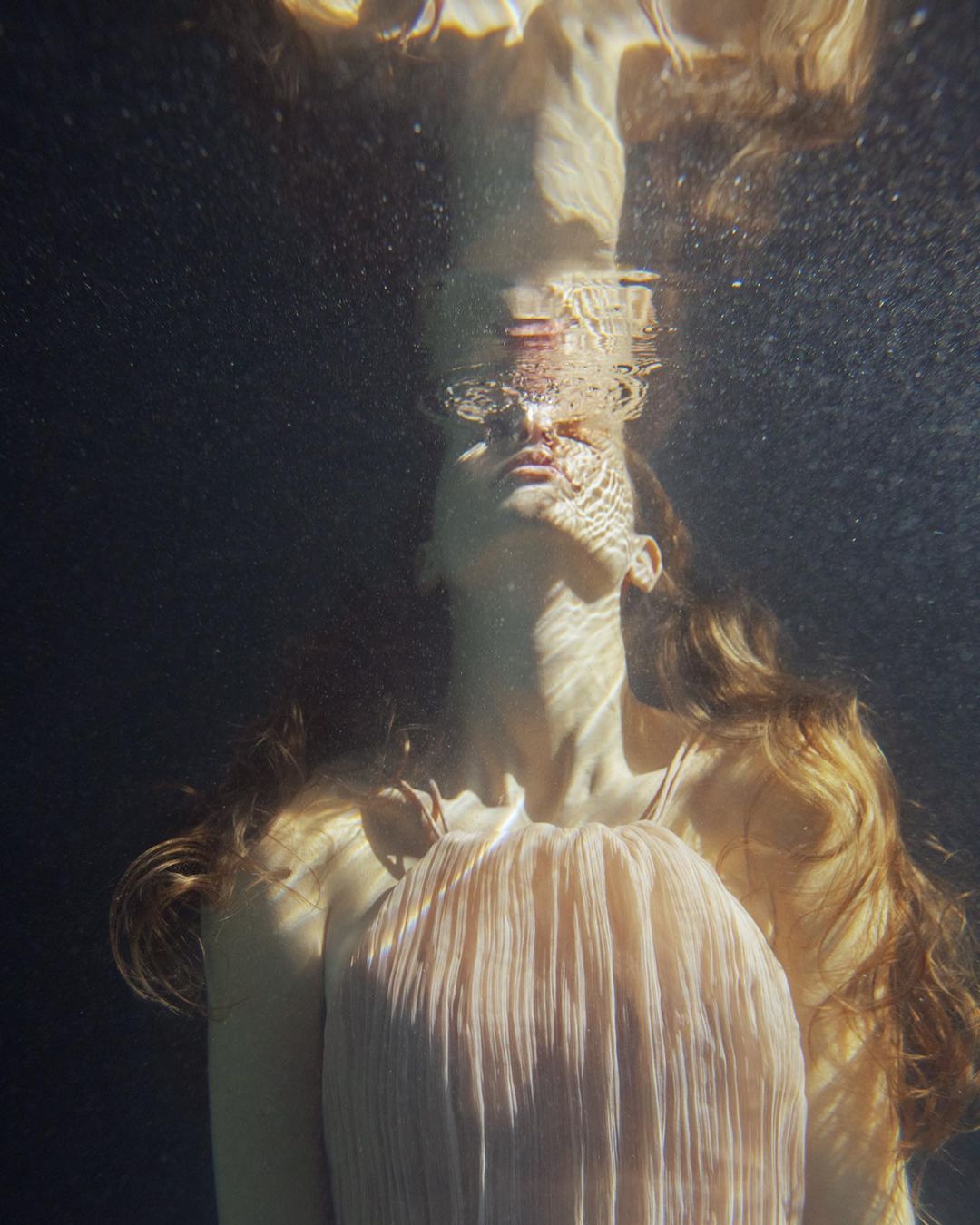 Underwater portrait of a woman with long wavy hair and a flowing dress looking up towards the water's surface with sunlit patterns.