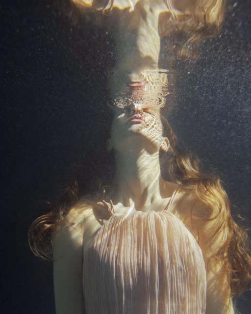 Underwater portrait of a woman with long wavy hair and a flowing dress looking up towards the water's surface with sunlit patterns.