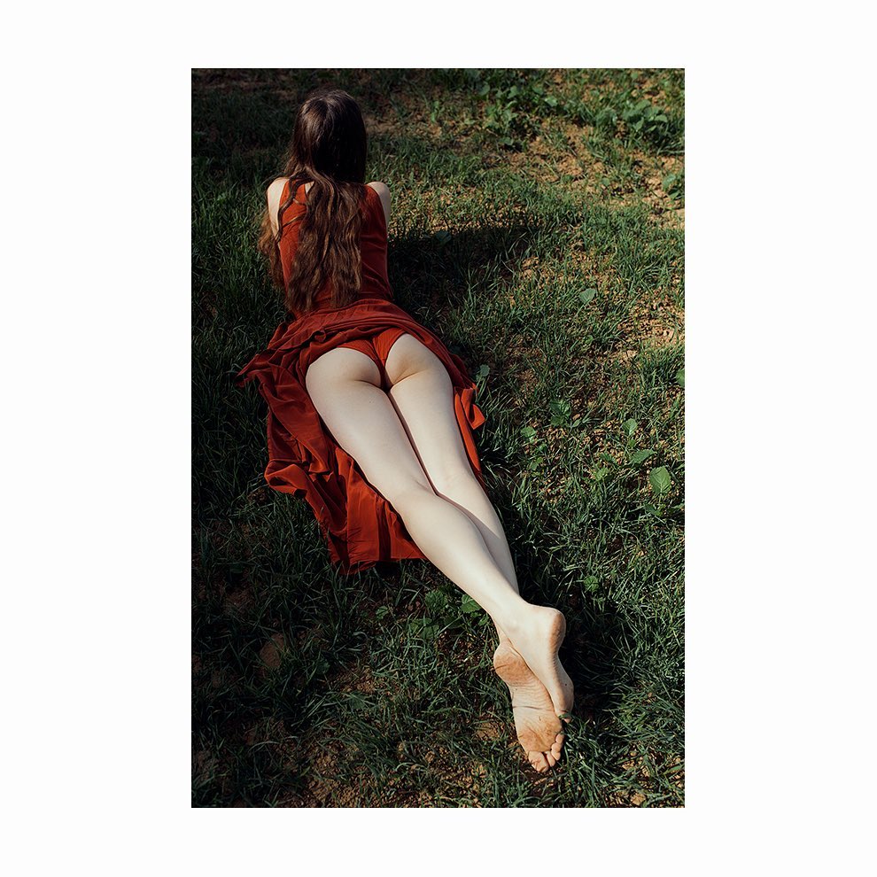 Barefoot woman lying on grass in a red dress. Back view of a female model outdoors in a grassy environment wearing a flowing red dress.