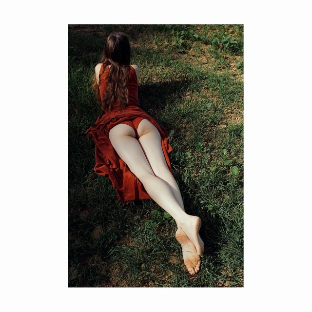Barefoot woman lying on grass in a red dress. Back view of a female model outdoors in a grassy environment wearing a flowing red dress.