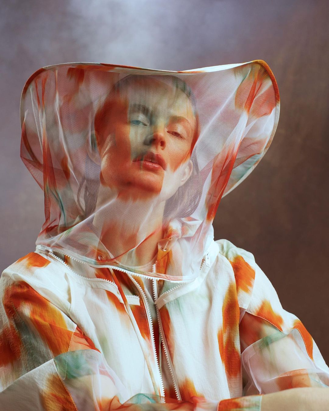 Close-up of a woman in a painterly-printed hooded jacket, with a semi-transparent veil over her head, against a muted brown backdrop.