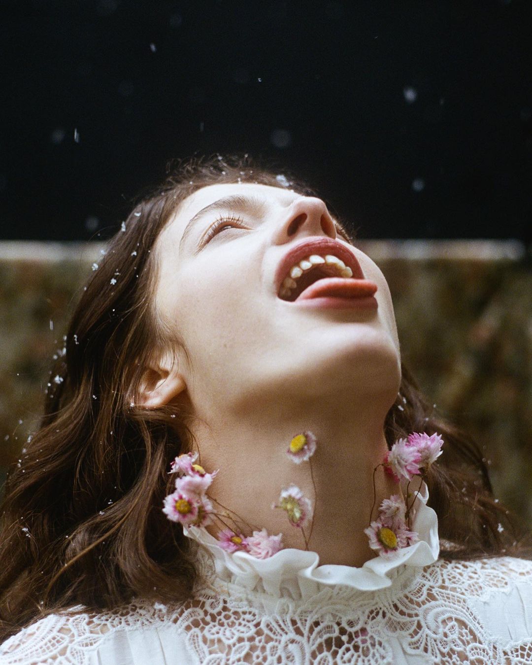 A woman lies on her back with her mouth open, sticking her tongue out. She is surrounded by small flowers and there are dark background.